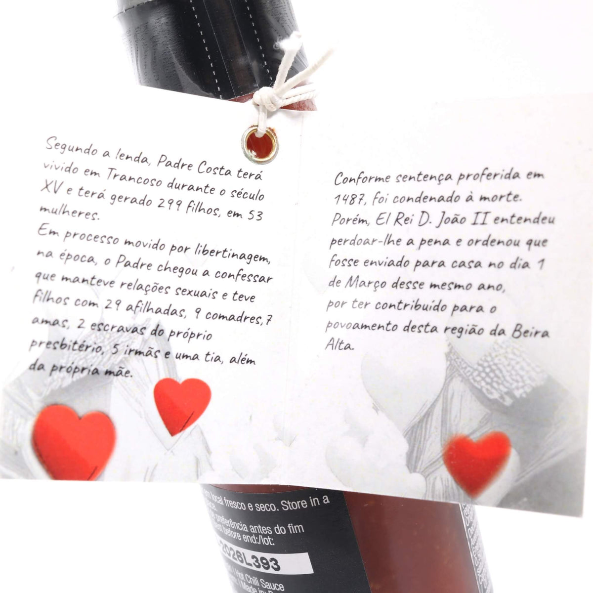 A close-up of a white printed card or label attached to a black bottle, featuring Portuguese text, two red hearts, and a small gold heart charm tied with string. Part of the bottle label is visible at the bottom.