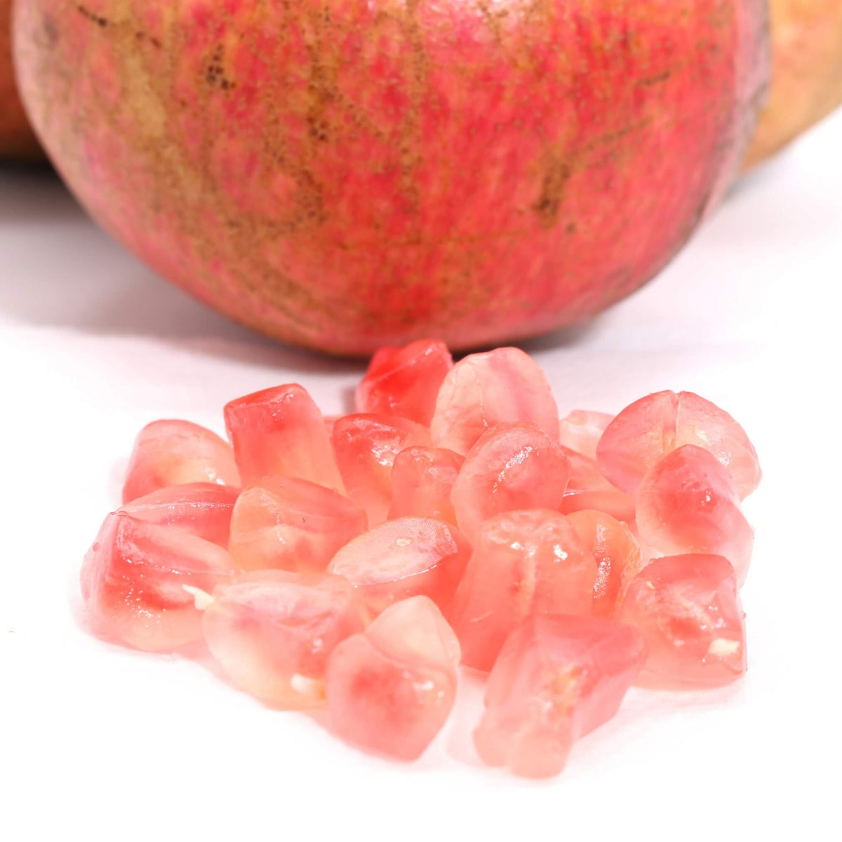 A close-up of loose pinkish-red pomegranate seeds in the foreground with whole pomegranate fruits blurred in the background on a white surface.